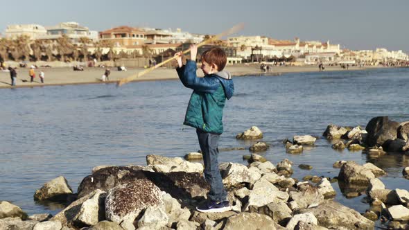 the Boy Spins a Stick with His Hands on the Rocks By the Sea by partem435