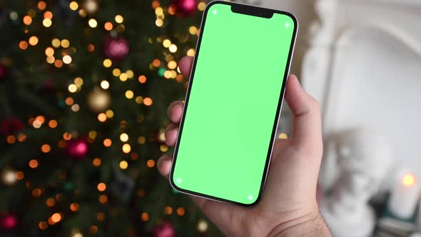Man holding modern smartphone with green screen chromakey near Christmas tree lights on background