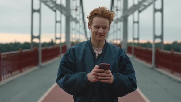 Smiling Man Walking on Bridge with Smartphone in Hands alt