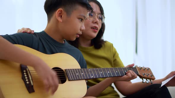 Asian mother embraces son, Asian boy playing guitar and mother embrace on the sofa alt