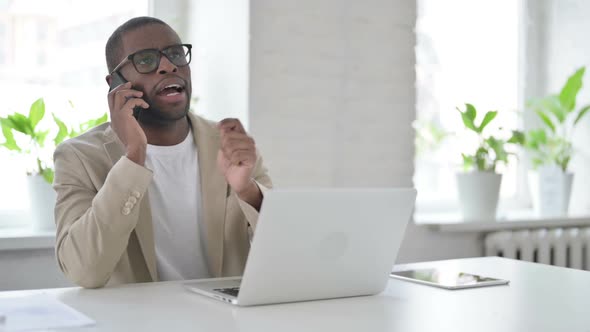 African Man Talking on Smartphone While Using Laptop in Office alt