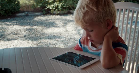 Little Tired Boy Learns Remotely From Home on a Tablet Sitting on the Terrace alt