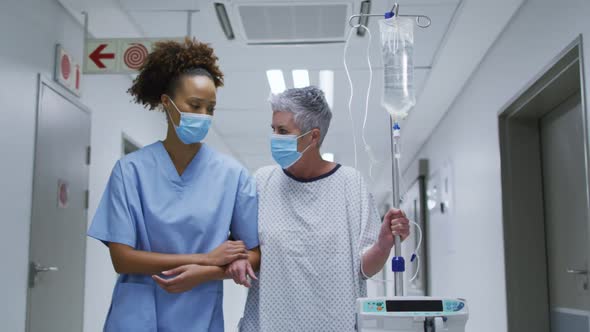 Female diverse doctor and patient wearing face masks walking through hospital corridor alt