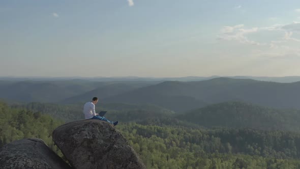 Aerial View of a Young Man Working on a Laptop Sitting on Top of a Rock Mountain in the Forest