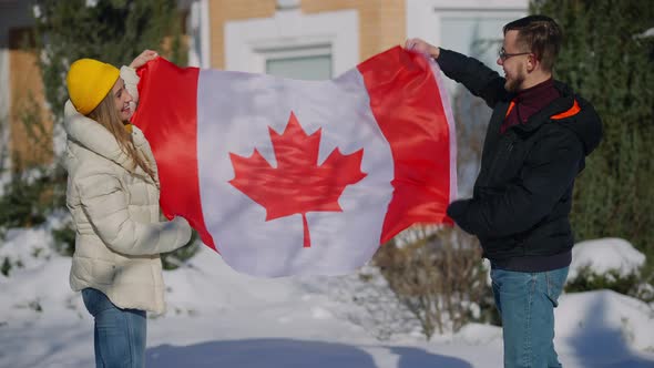 Young Happy Canadians Posing with National Flag Fluttering in Hands Outdoors on Sunny Winter alt