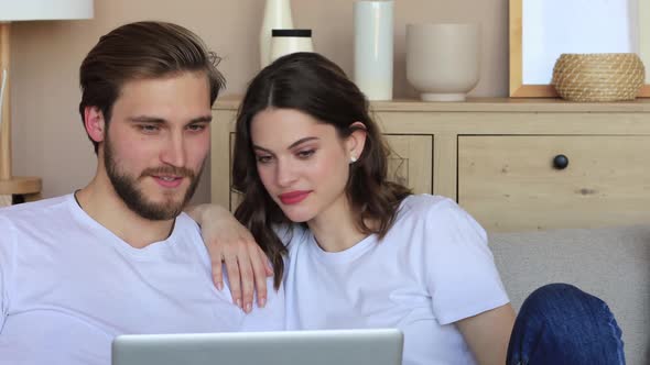Happy young couple sitting on couch at living room and looking at camera during video chat alt