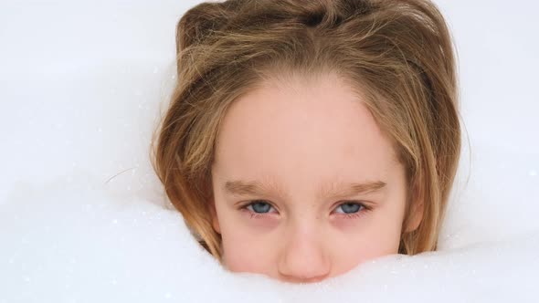 Portrait of Cheerful Little Girl Lying in Bath with Thick Soap Foam and Looking in Camera