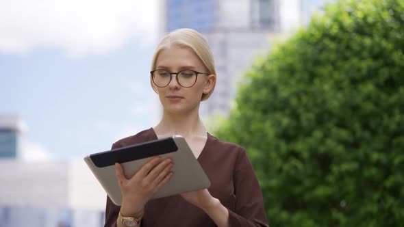 A Young Blonde Woman in Glasses Scrolls the Tablet with Interest in the Street alt