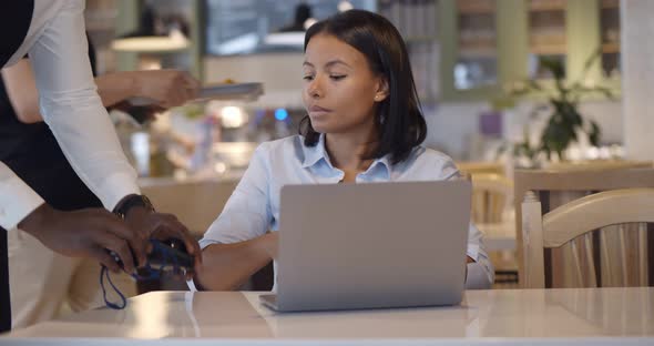 Afro Businesswoman Paying with Credit Card Sitting at Table with Laptop in Cafe alt