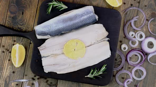 Fillet of Salted Herring with Onions Lemon and Rosemary on Dark Cutting Board Rotating alt