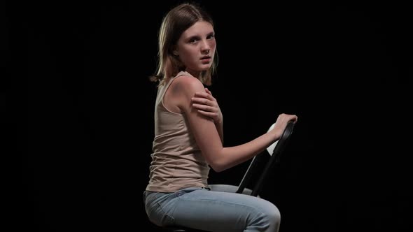 Side View Beaten Teenage Girl Showing Bruise on Shoulder Sitting on Chair Looking at Camera alt