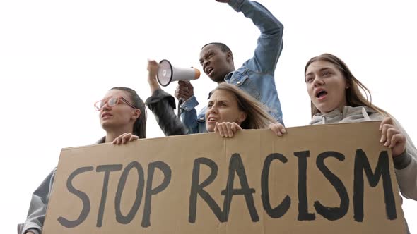 Group of Young People Holds a STOP RACISM Poster. Dark-Skinned Young Protester Shouts Slogans Into a alt