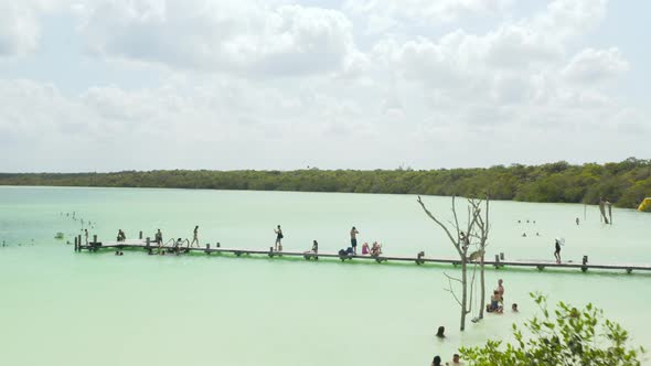 Fly Over People Relaxing at Water Sitting at Long Wooden Pier or Bathing in Natural Swimming Pool alt
