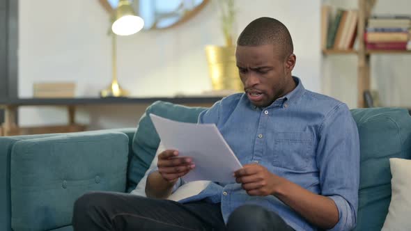 Young African Man with Documents Reacting to Loss on Sofa alt