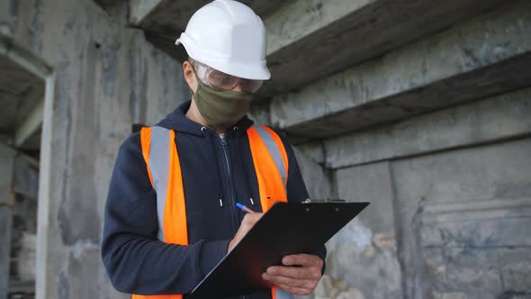 A Male Foreman in a Construction Helmet and Protective Antiviral Mask at a Construction Site Makes