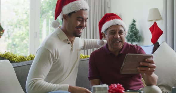 Cheerful biracial son with senior father in santa hats making video call on tablet pc in christmas alt