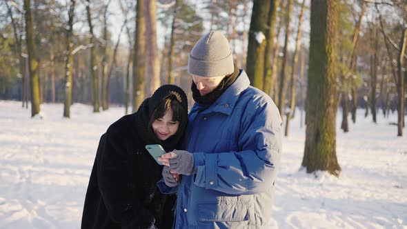 Young Couple in a Winter Park Watching Their Selfies in a Smartphone alt