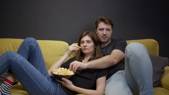 Attractive Young Couple Sitting on Sofa with Popcorn Watching Movie on TV