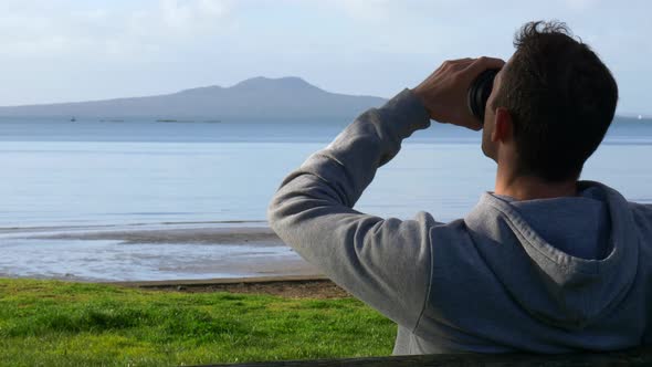 Young caucasian man drinking coffee sitting on a bench at the beach  looking at Rangitoto island in alt