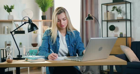 Blond Young Woman Working with Laptop and Doing Needed Notes Into Papers in Home Office alt