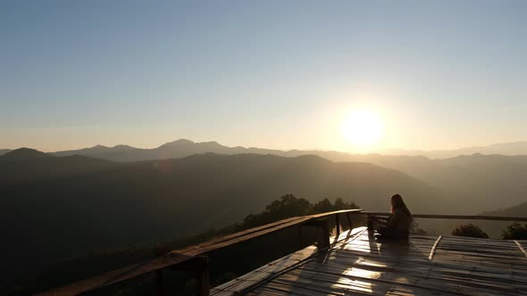 A female sitting and playing guitar on wooden balcony with a beautiful mountain view before sunset