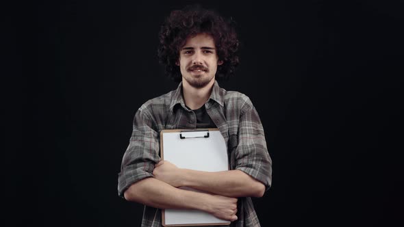 The Handsome Boy Hipster with Curly Hair Displays a Wide Smile Holds a Planner and a Pen in His Arms alt