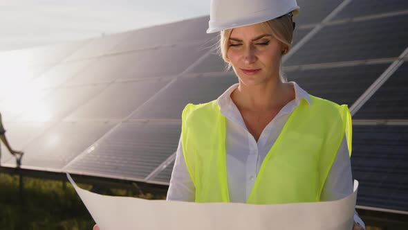 Beautiful Female Solar Engineer Standing Near the Panels with a Drawing on Paper and a Young alt
