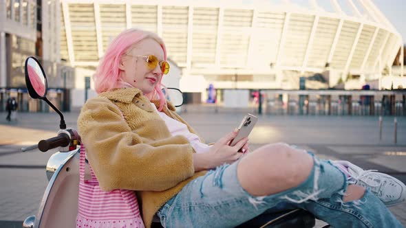 Young Stylish Woman with Pink Hair Relaxing on Scooter and Web Surfing Social Media on Smartphone alt