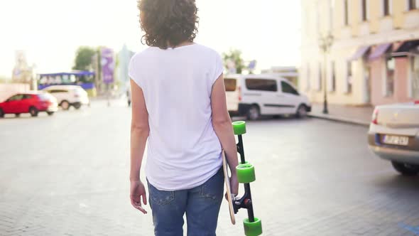Back View of a Woman Walking in the Old City Street Holding Her Trendy Stylish Longboard with alt