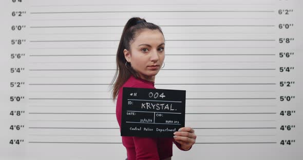 Mugshot of Arrested Millennial Woman Turning Head and Looking to Camera While Standing Aside alt