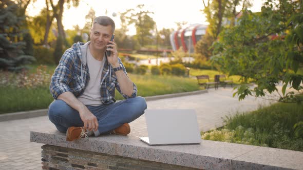 A Male Freelancer Working on a Laptop in the Park Communicates on the Phone Discussing Work Issues