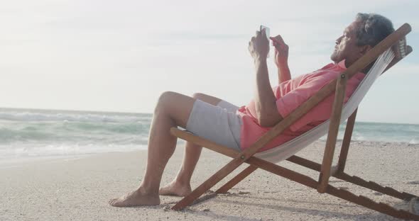 Latin senior hispanic man relaxing on sunbed on beach at sunset, using smartphone alt