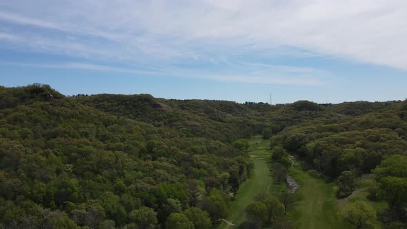 Drone view over lush green tree filled valley with golf course at the bottom. Blue sky with clouds