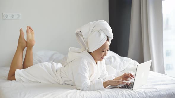 Cute Asian Woman Dressed in a Bathrobe and a Towel on Her Head Working on a Laptop Lying in Bed alt