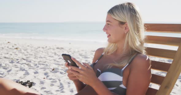 Caucasian woman sitting on a sunbed and using her smartphone on the beach alt