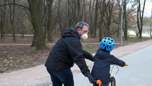 A father teaches his son to ride a Bicycle in a city Park. they are wearing protective helmets