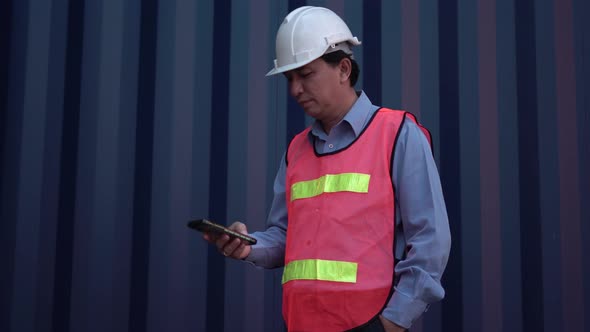 Asian man in hard hat standing with using smartphone at container cargo site
