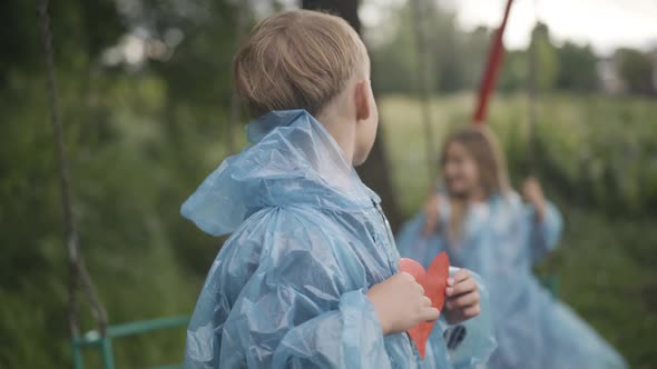 Confident Caucasian Boy with Paper Heart Looking Back at Girl Swinging on Swings and Smiling at alt