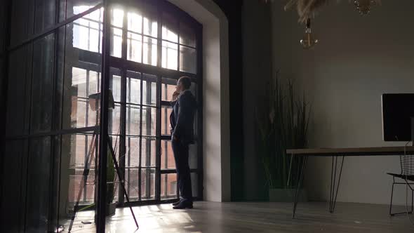 in a office a businessman speaks on the phone while standing by the window alt
