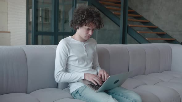 Concentrated Curly Guy Typing on Laptop While Sitting on Sofa in Fashionable Apartment alt