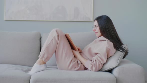 Young caucasian woman with glasses sitting on sofa in cozy living room, smiling and reading book.