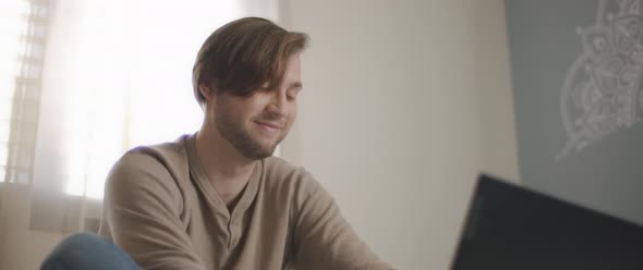 A close up of a young man smiling while sitting in bed and working on a computer.  alt