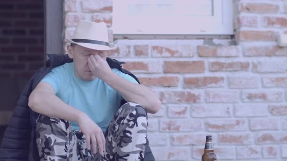 A Young Man in a Hat Sits on the Porch of the House Next to a Bottle of Beer alt