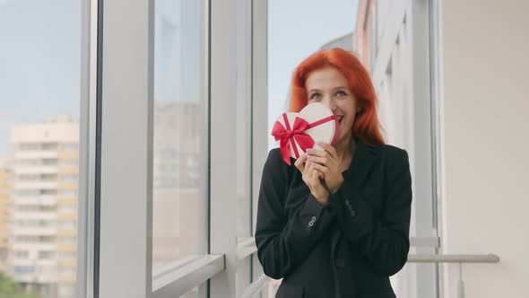 The Woman Received a Gift Box in the Form of a Heart. Happy Woman with Red Hair Rejoices at the Gift alt
