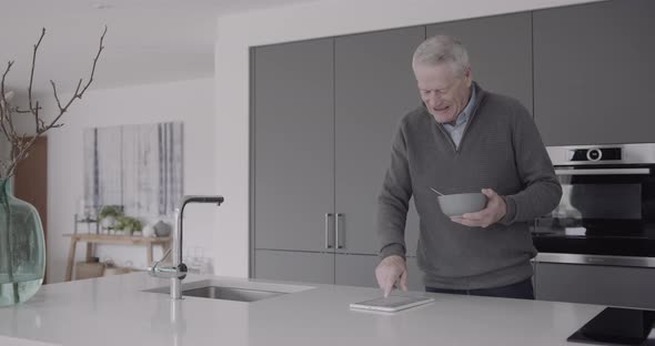 Senior man eating healthy breakfast in kitchen and looks on digital tablet