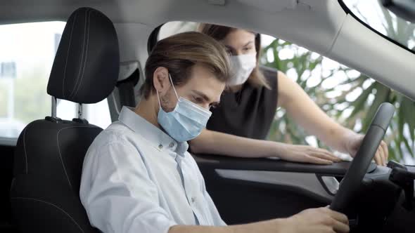 Young Man in Covid-19 Face Mask Sitting on Driver's Seat in Car As Blurred Woman Talking To Buyer in alt