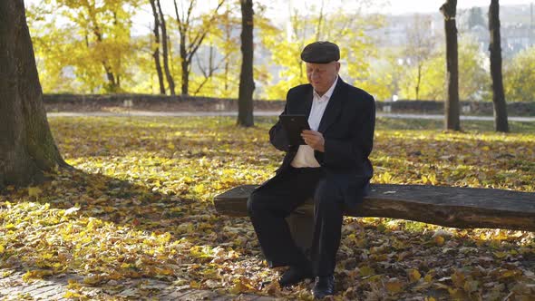 Happy Senior Man Browses the Tablet Screen When Resting on Bench in Autumn Park alt