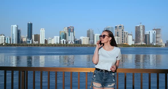 Young Beautiful Girl Standing on the Waterfront Against the City Talking on the Phone alt