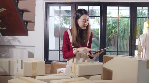 Asian female entrepreneur checking customer order on digital tablet while packing boxes