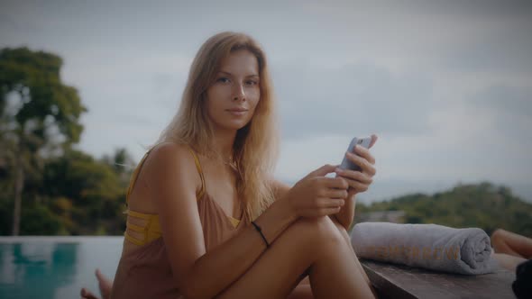A Blonde Woman Uses Her Mobile Phone While Lounging in the Sun By the Pool on Vacation alt